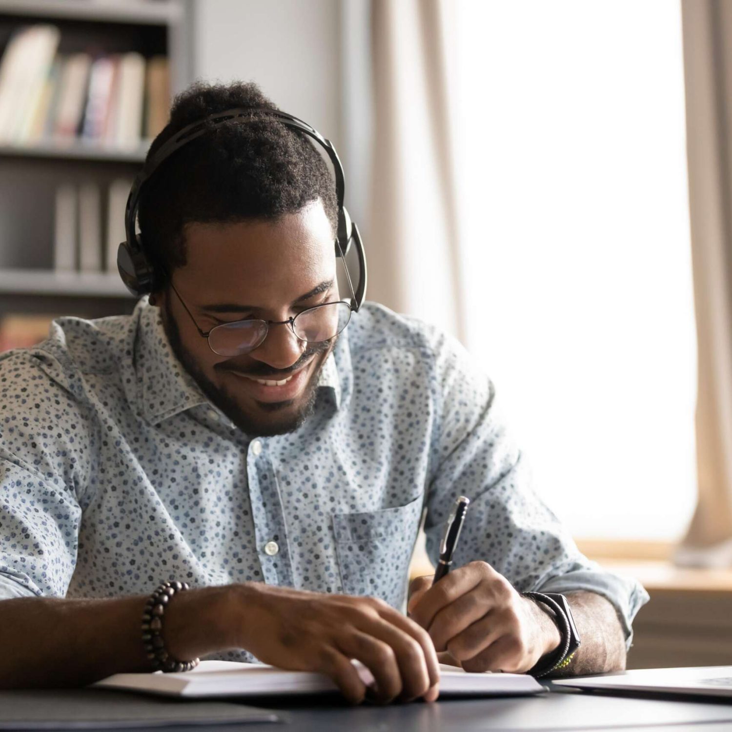 smiling-motivated-african-american-student-wearing-headphones-listening-lecture-writing-notes-notebook-sitting-work-desk-learning-language-online-watching-webinar-elearning-concept-min